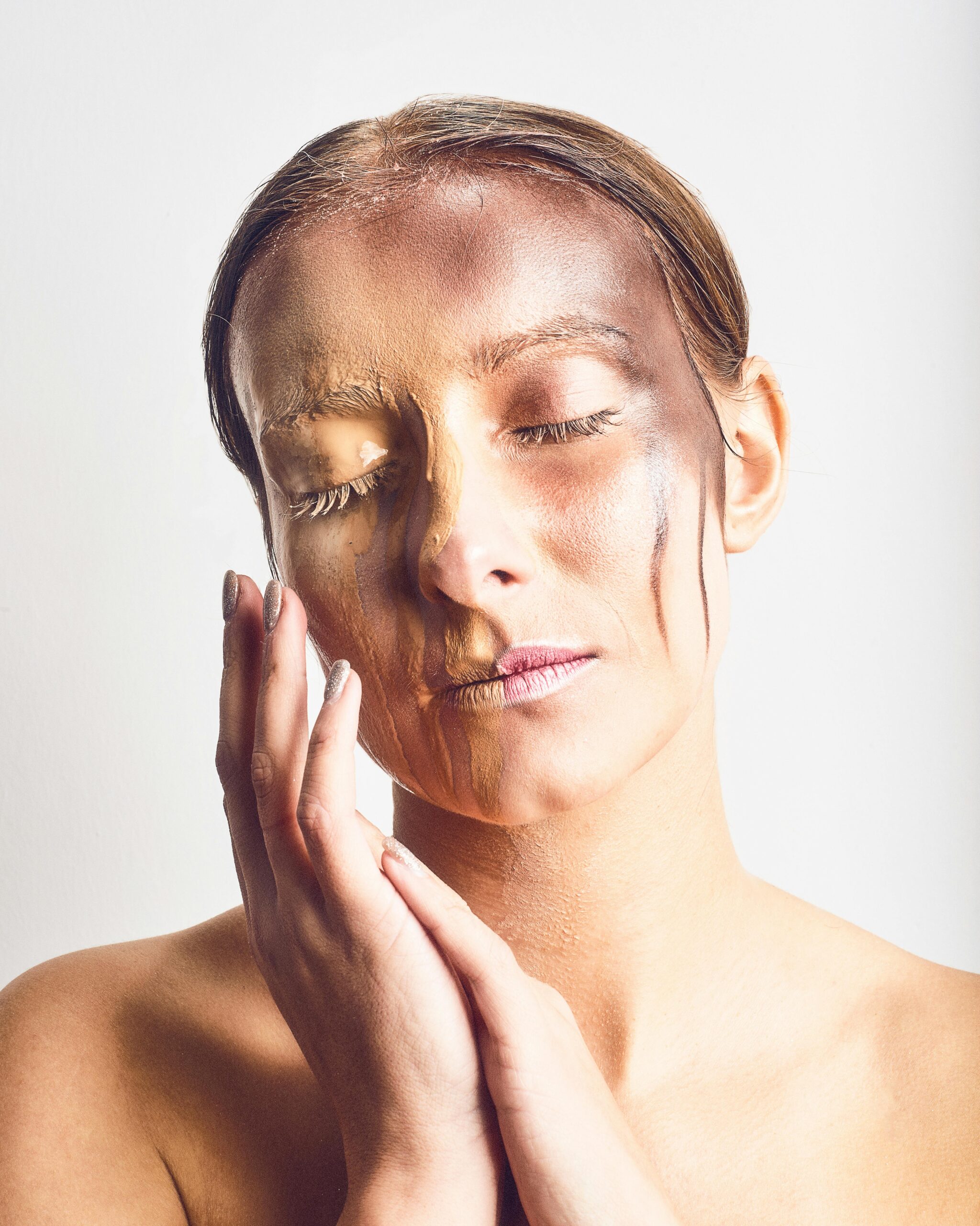 A woman with closed eyes and creative makeup blending multiple skin tones on a neutral background.