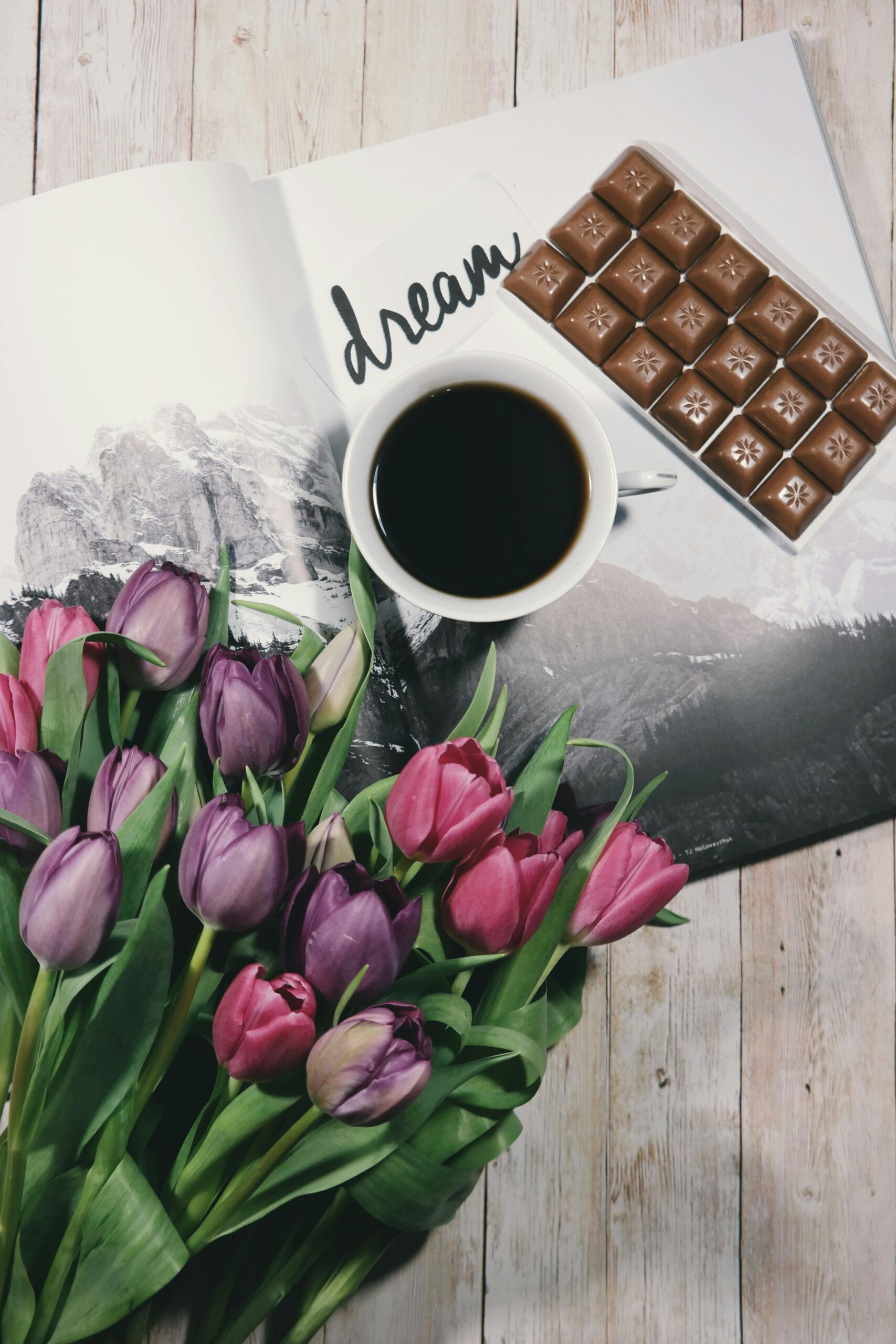 A warm setup featuring a cup of black coffee, colorful tulips, and chocolate on a wooden table.