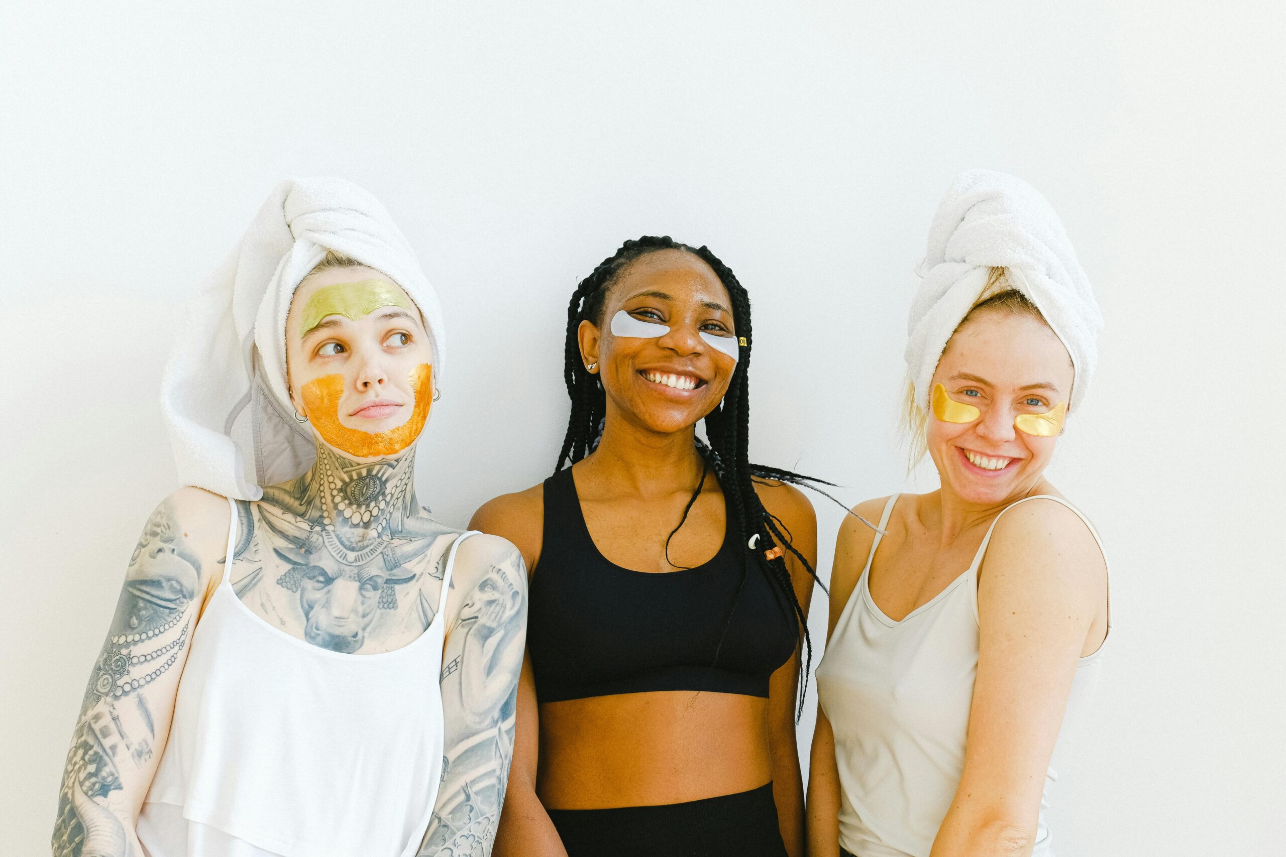 Happy women with masks and towel smiling and demonstrating beauty procedure while standing near white background together during weekend and enjoying