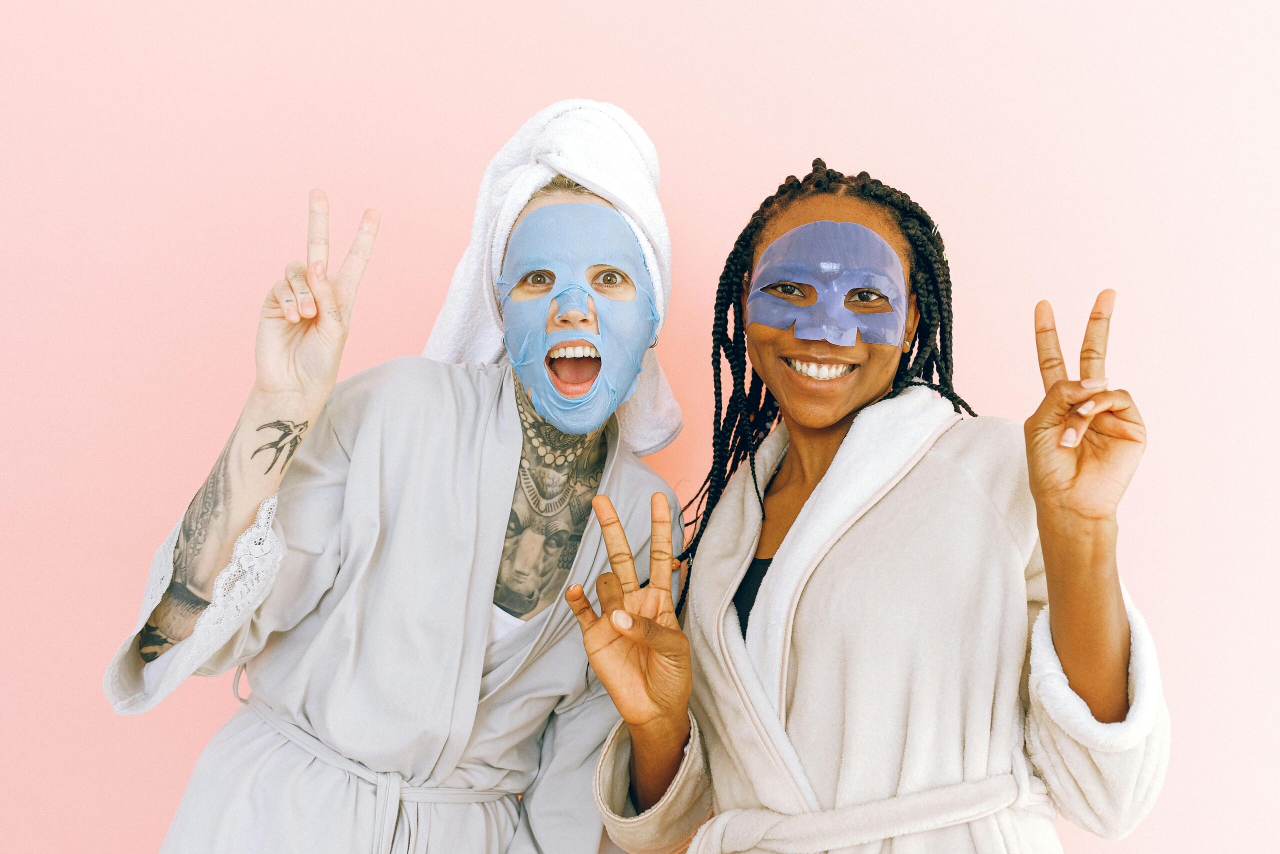 Two women having fun with skincare face masks, flashing peace signs in bathrobes.