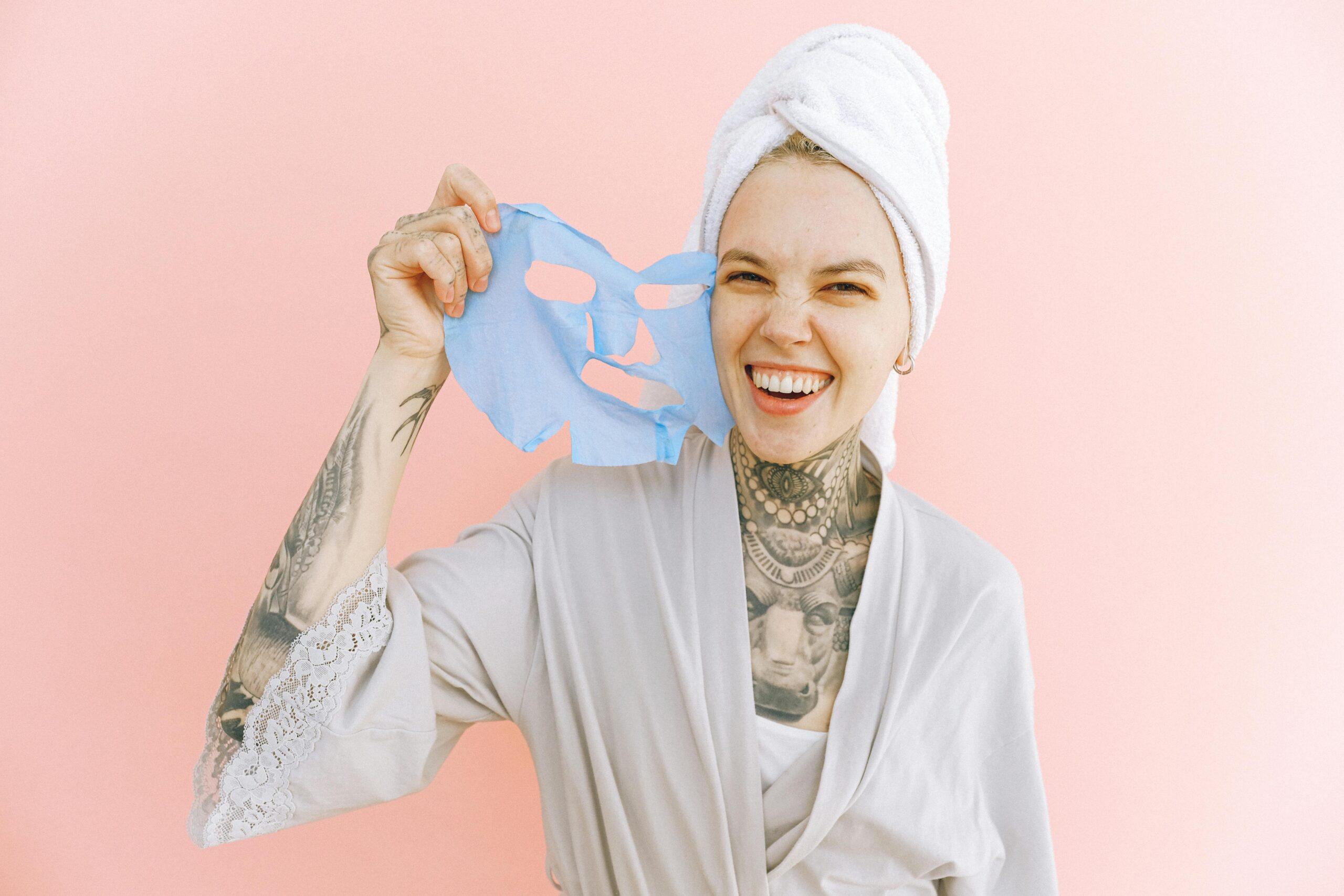 Happy woman holding a blue face mask, wrapped in a towel and smiling brightly.