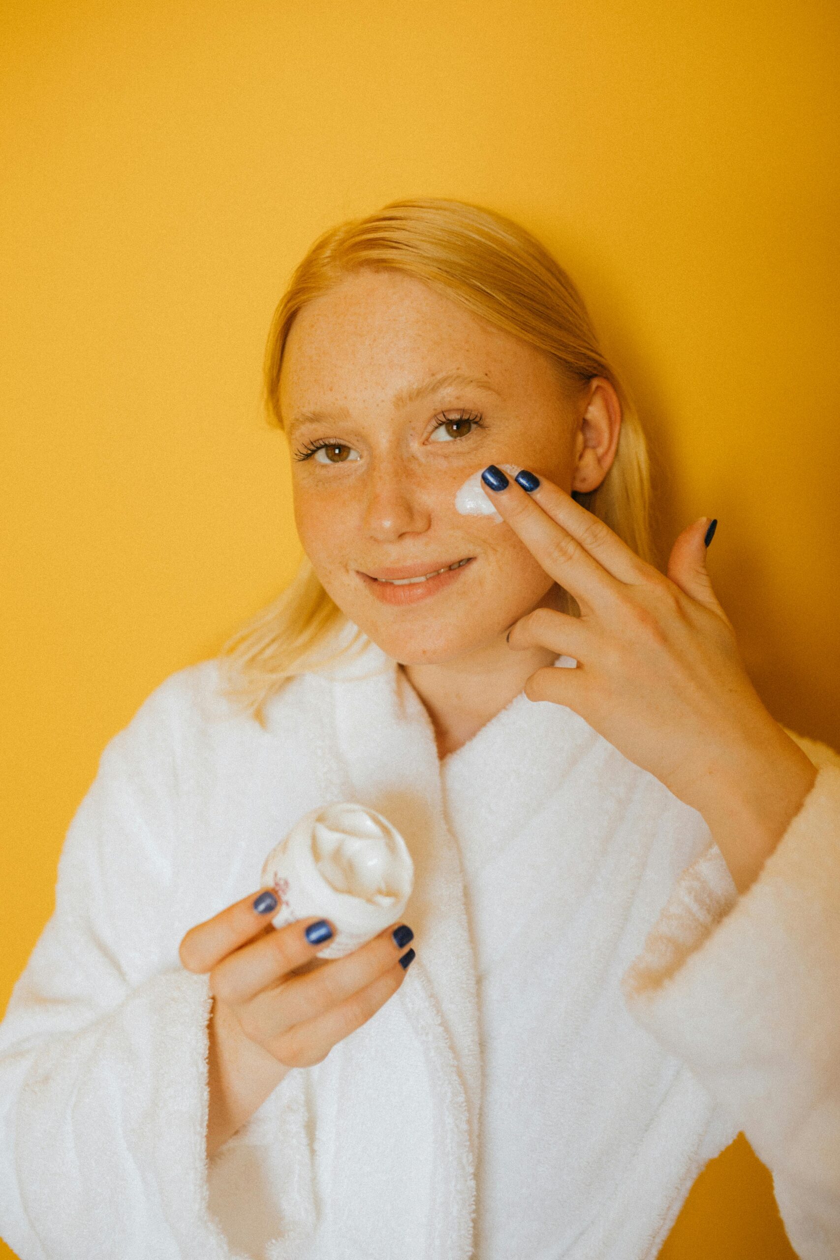 Woman in a white bathrobe applying skincare cream on a vibrant yellow background.