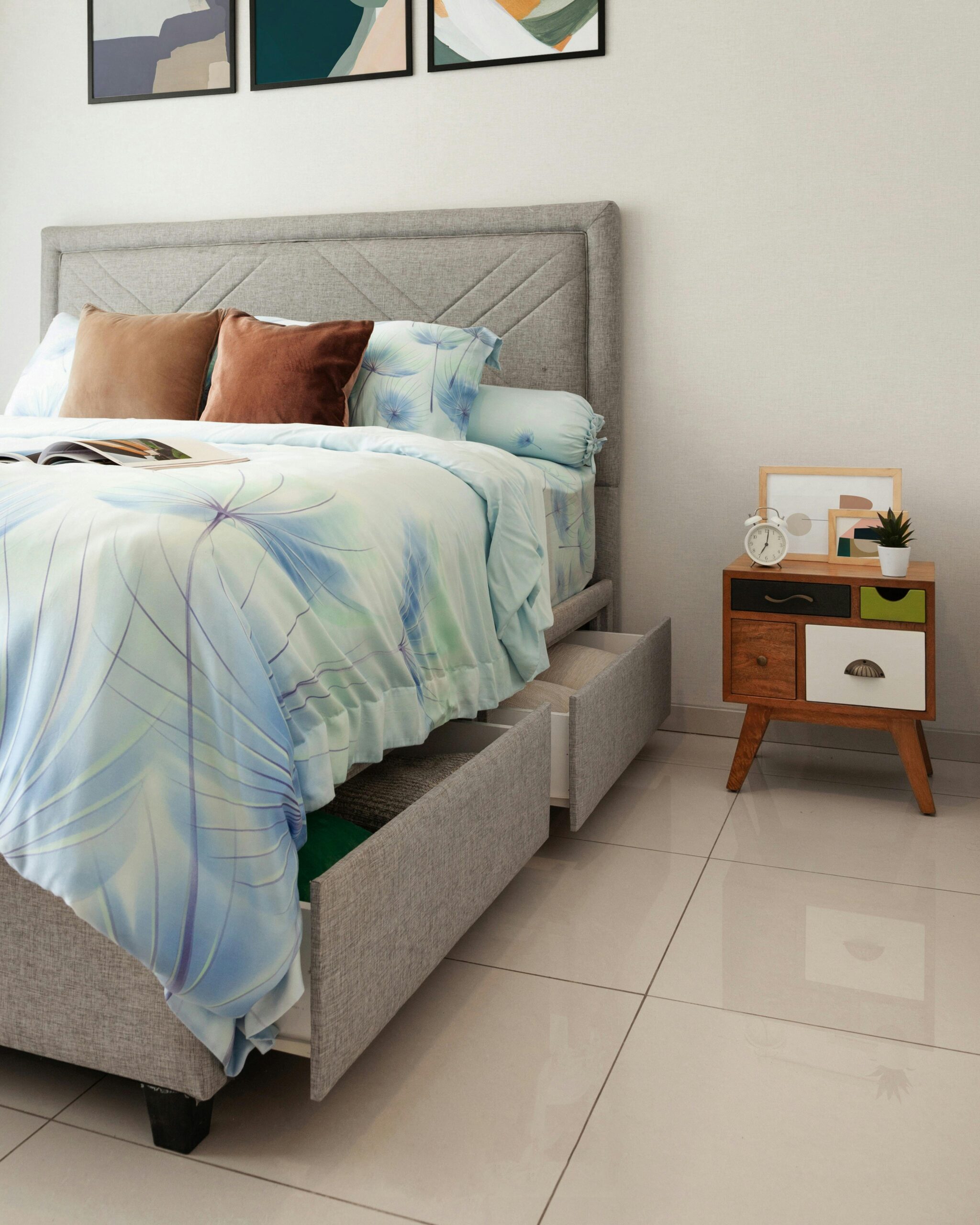 A modern, stylish bedroom featuring a grey upholstered bed with drawers, abstract artwork, and a mid-century side table.
