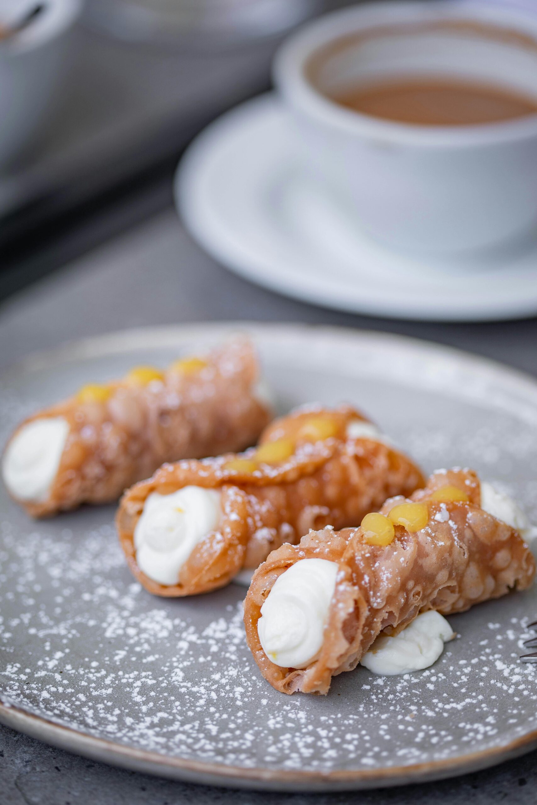 Three cannoli filled with cream paired with a cup of coffee on a grey plate.