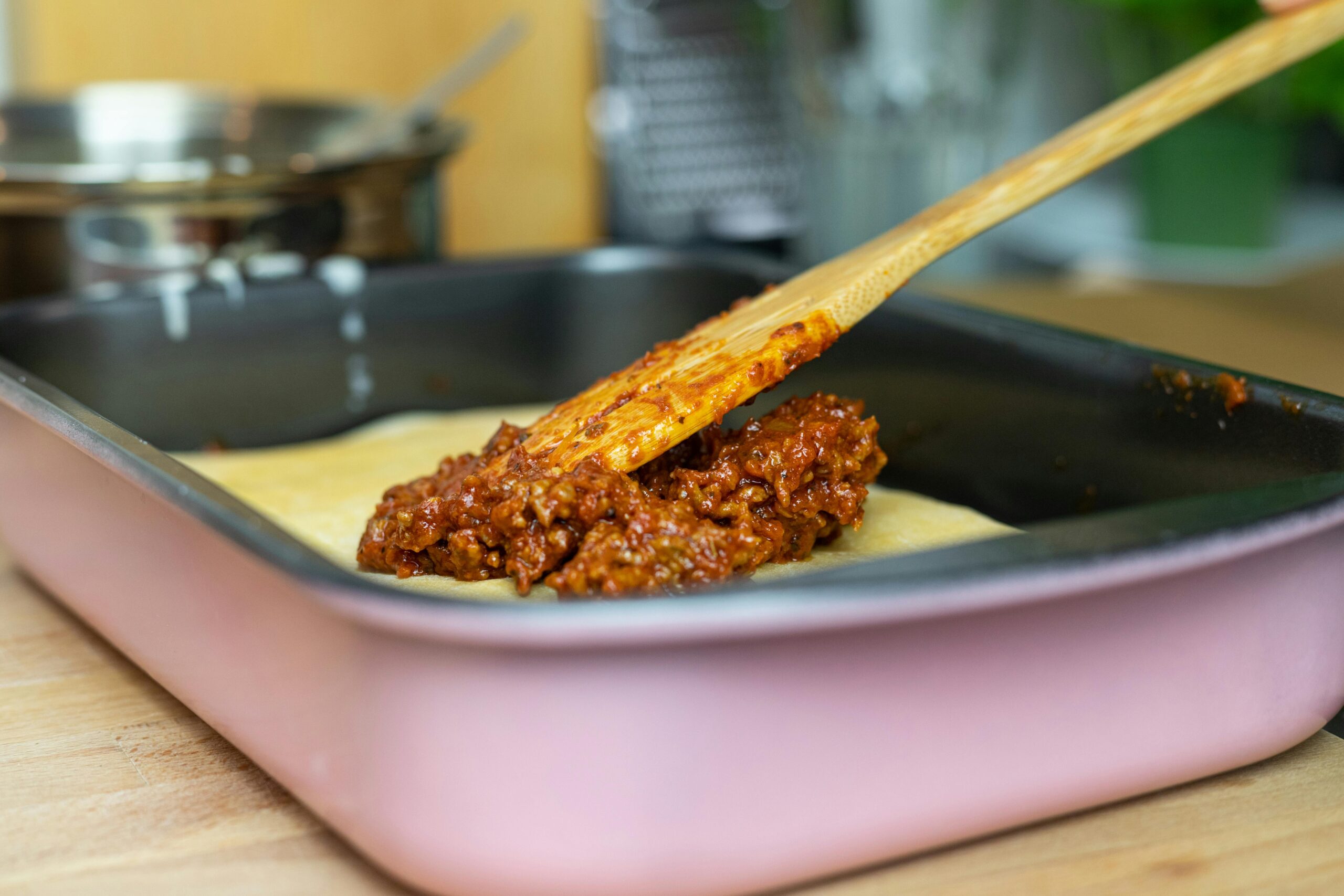 Close-up of layering meat sauce on lasagna in a pan in a kitchen setting.