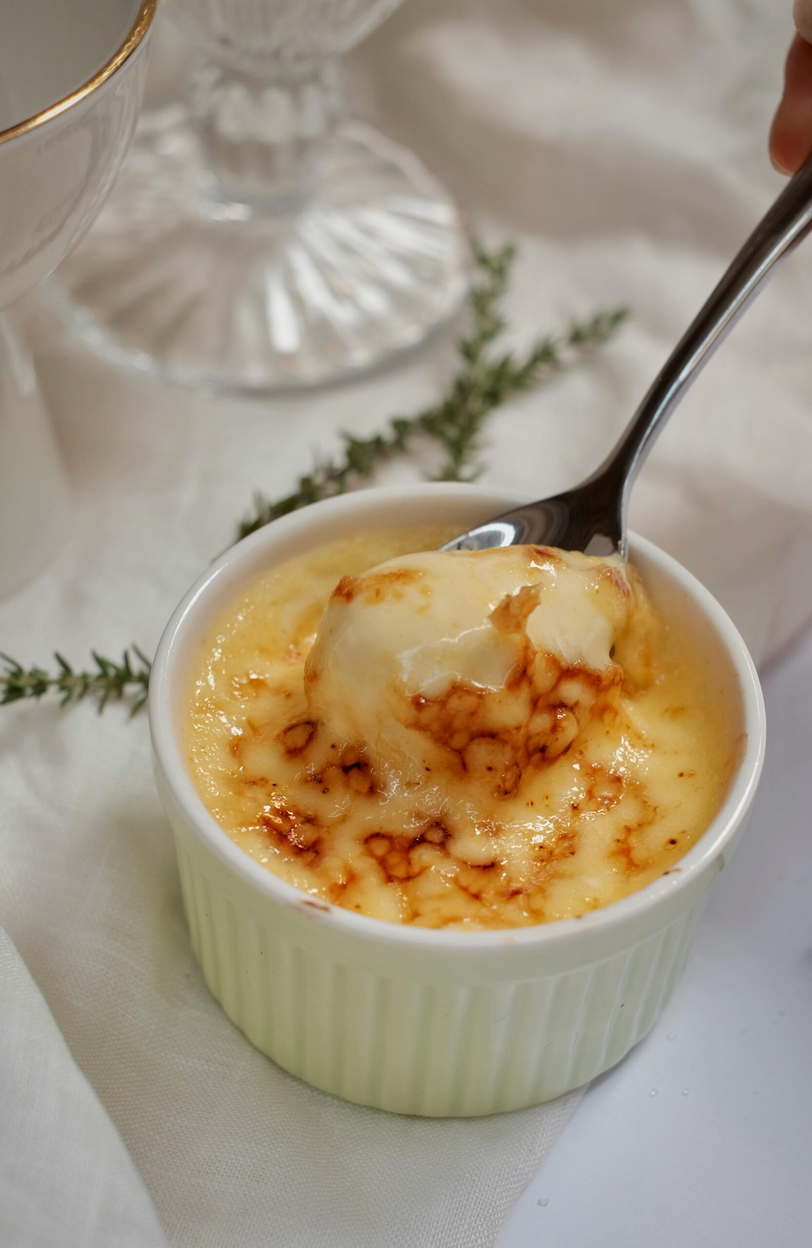 Close-up of a creamy crème brûlée dessert in white ramekin with caramelized top, garnished with herbs.