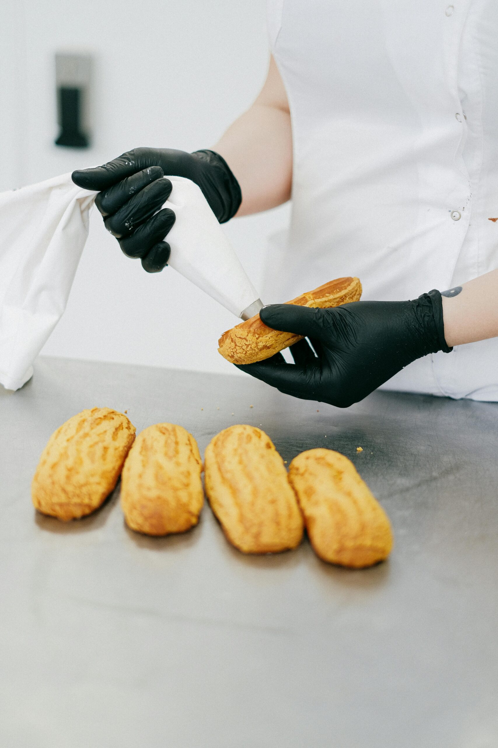 Pastry chef using piping bag to fill freshly baked eclairs on a stainless steel counter.