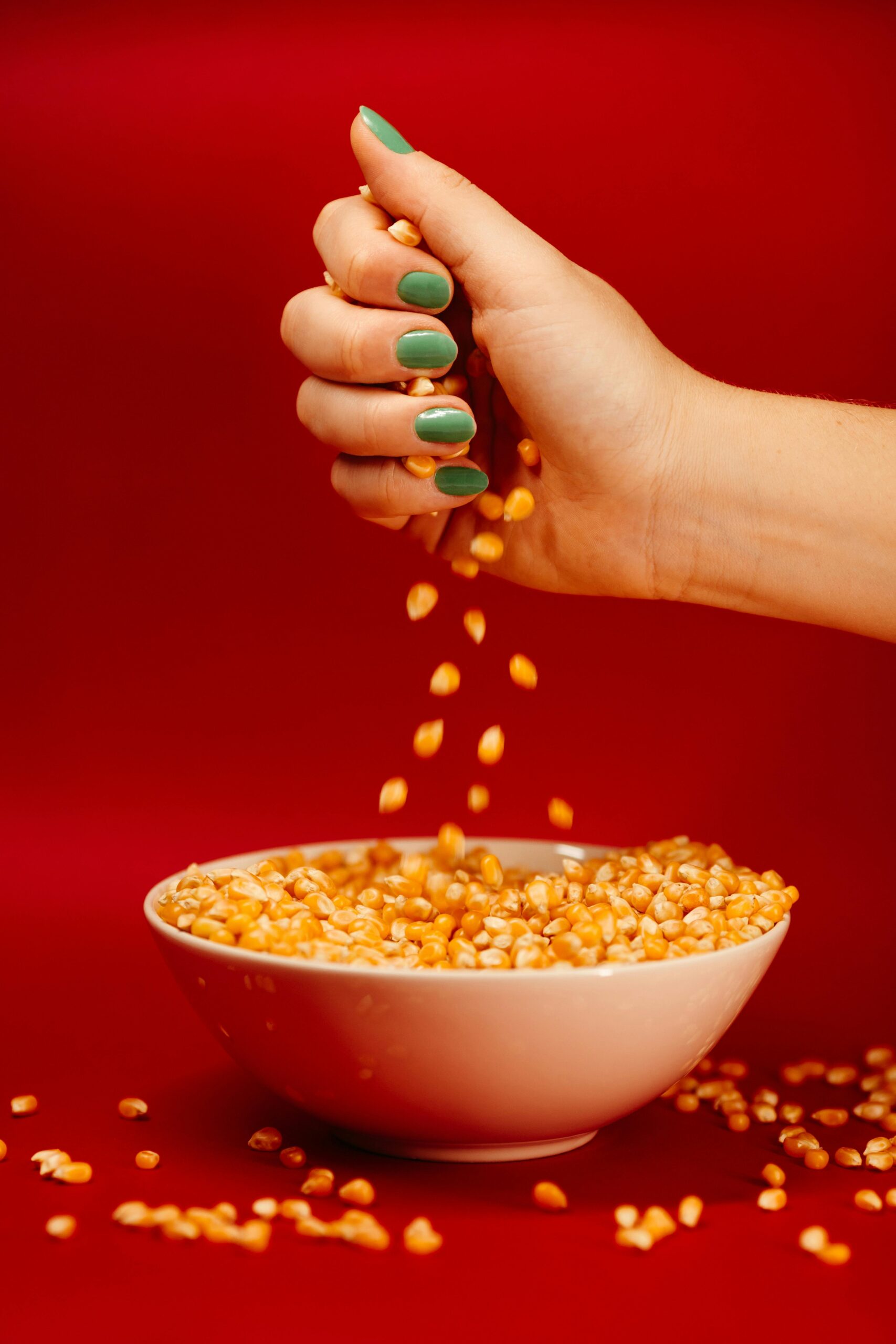 A woman's hand with green nails pours corn kernels into a white bowl against a red background.