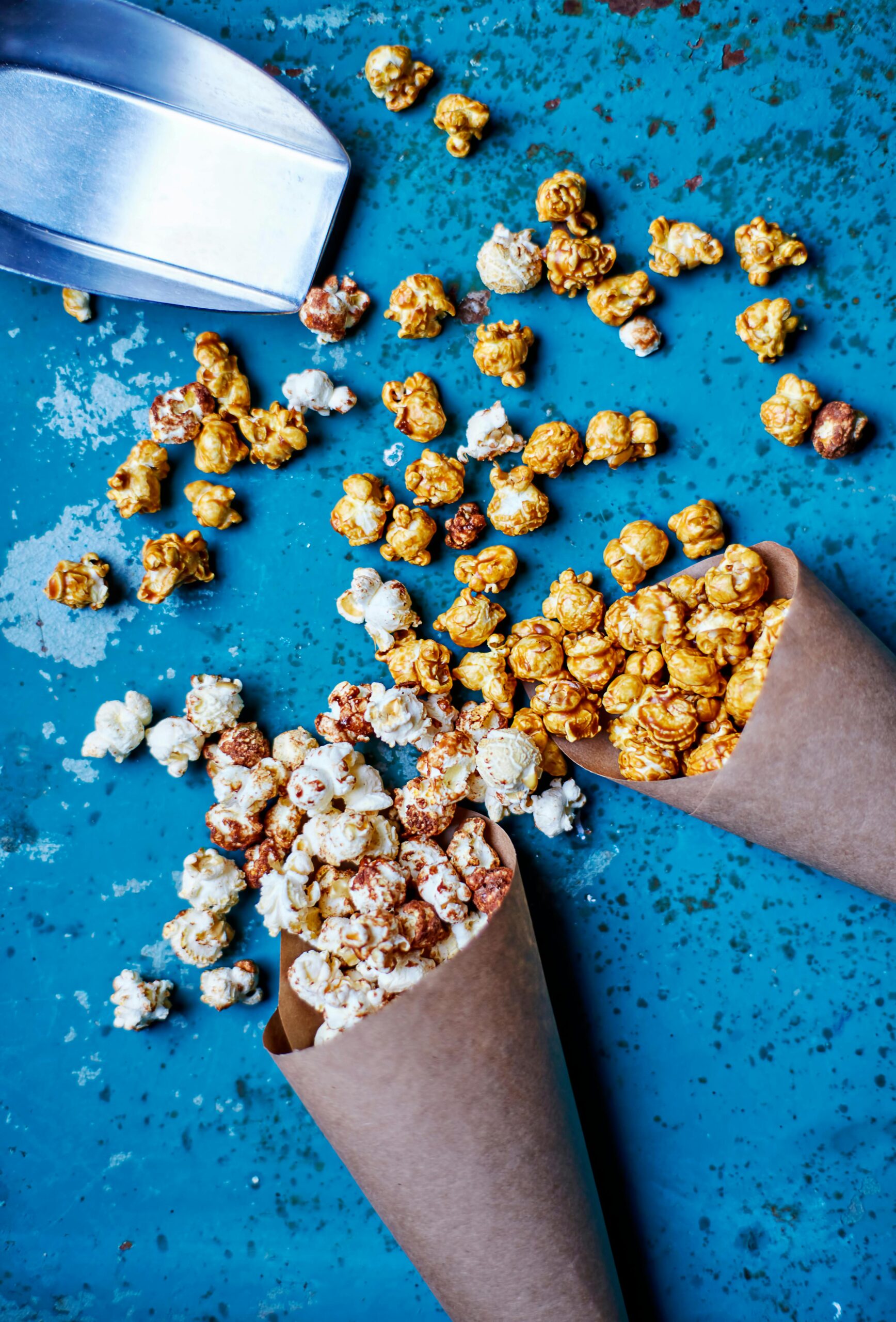 Gourmet popcorn scattered from paper cones on a blue surface with a metal scoop.