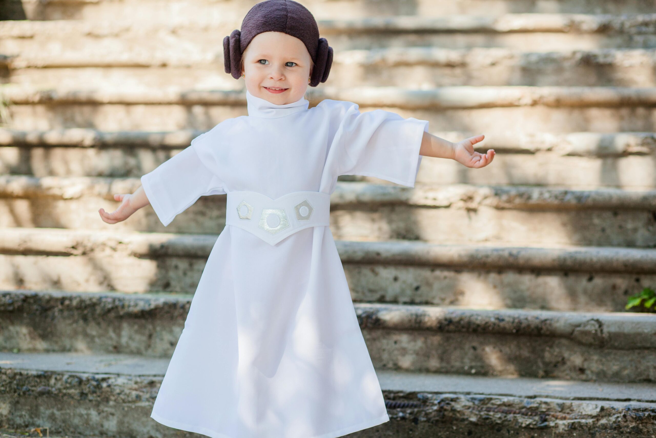 A little girl gleefully posing in a Princess Leia costume on outdoor steps during daytime.